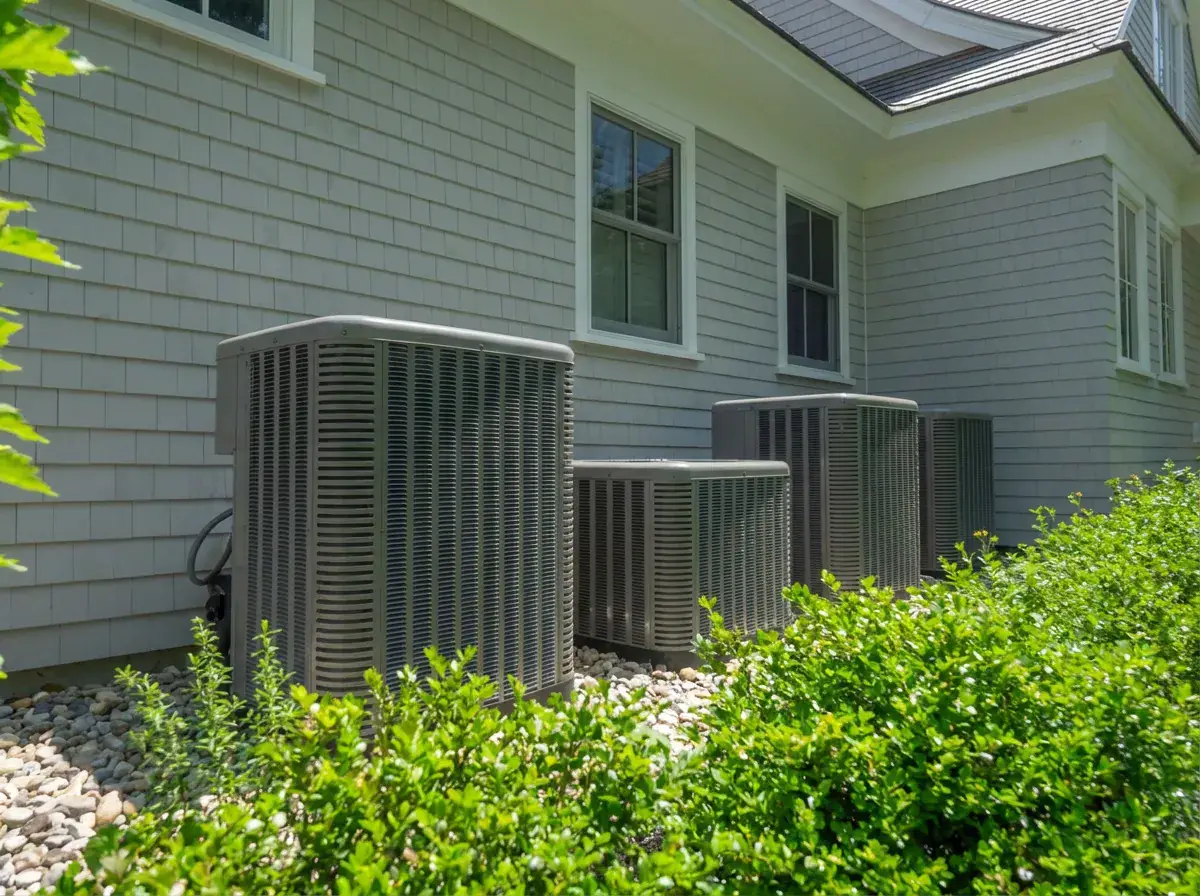 Three air conditioning condensers beside a grey shingled house, set back in a bed of shrubs