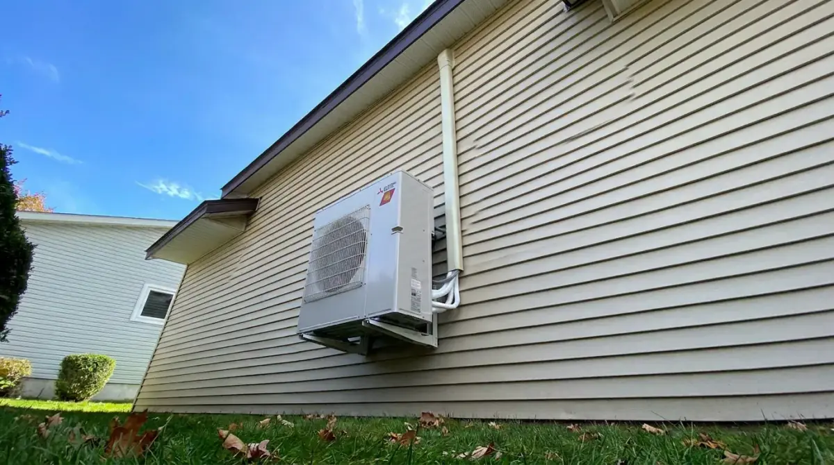 A mini-split outdoor unit mounted on the siding of a house with fallen autumn leaves on the lawn