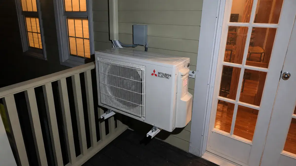 A Mitsubishi ductless mini-split outdoor unit mounted on brackets beside a lit porch door at night