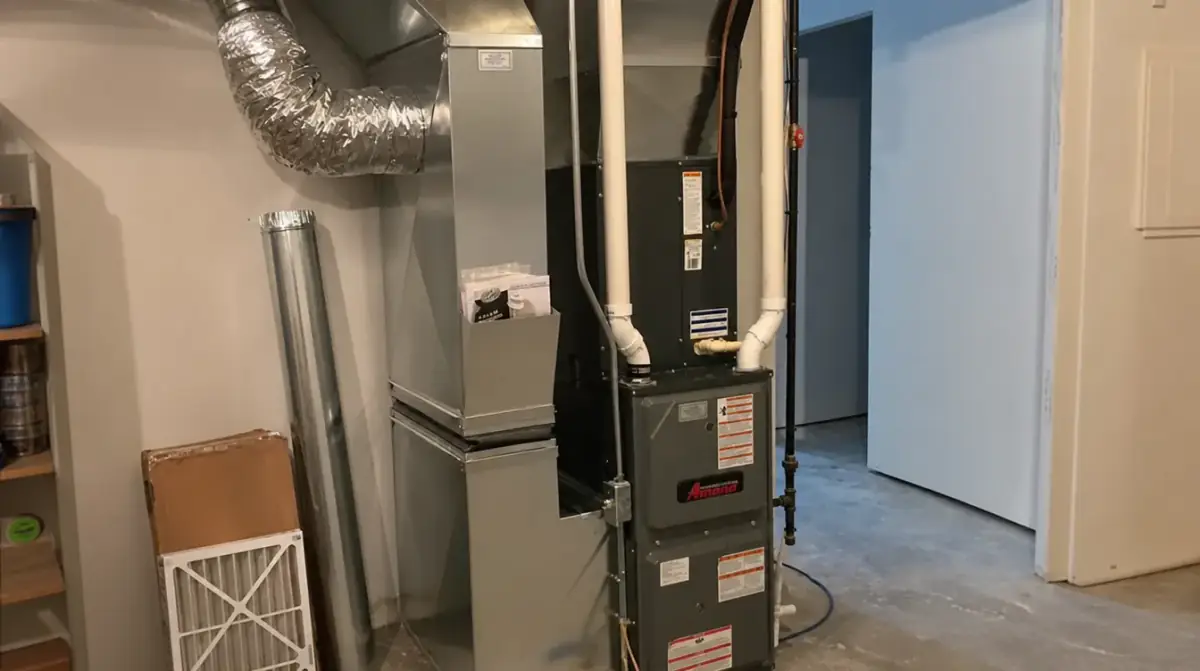 A basement furnace connected to insulated flexible duct, with a spare pleated air filter leaning against the wall beside it
