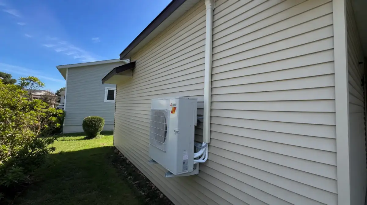 A mini-split heat pump outdoor unit mounted on the siding of a pale yellow house under a clear sky