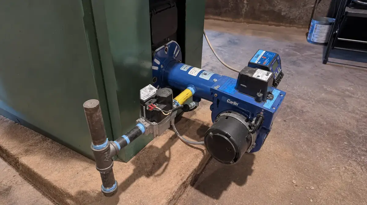 A blue burner unit bolted to the front of a green boiler, with the fuel line and controls attached